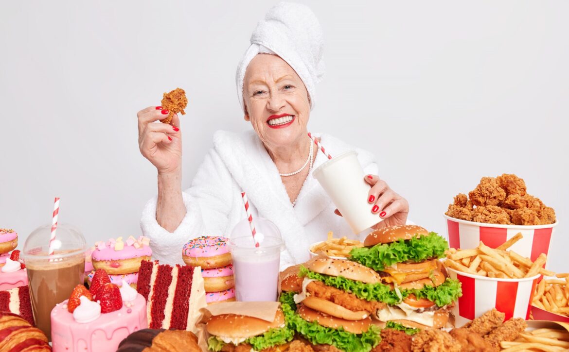 Unhealthy overeating lifestyle. Pleased old lady smiles positively drinks soda eats fast food suffers from gluttony bad eating habits has happy mood isolated over white background wears bathrobe